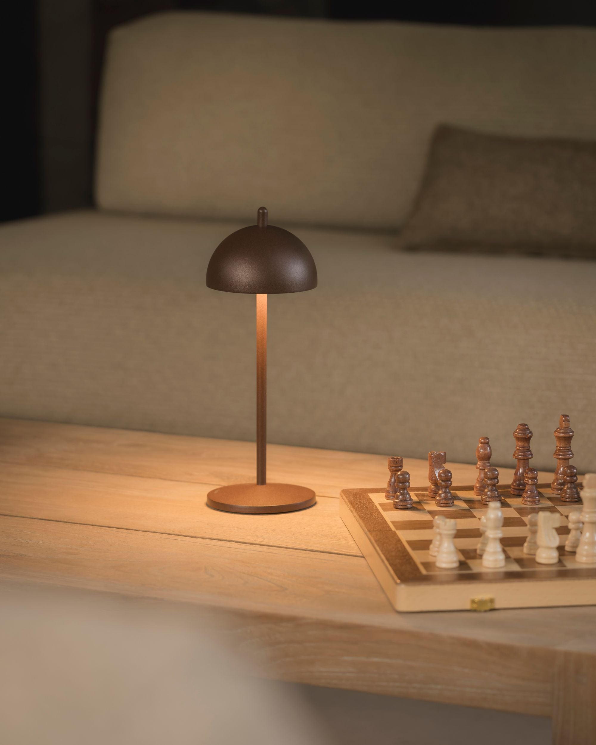 SAMOS cordless table lamp in Corten, casting warm light on an oak coffee table beside a wooden chess set
